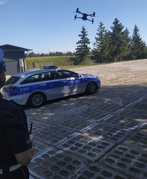 Radiowóz policyjny, dron policyjny i fragment sylwetki funkcjonariusza policji obsługującego drona